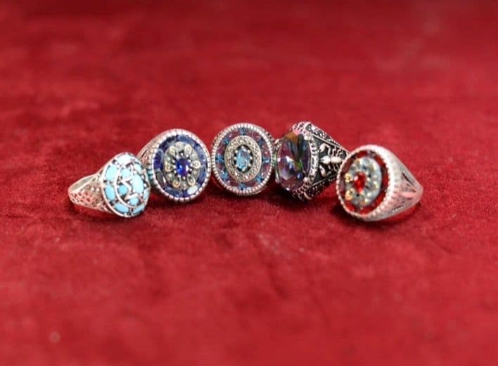 A collection of five ornate sterling silver-toned rings featuring a variety of semi-precious gemstones including turquoise, blue sapphires or lapis, a multi-color iridescent stone (likely mystic topaz or similar), and a ruby-red stone, each set with intricate filigree or decorative metalwork on a red velvet background.
