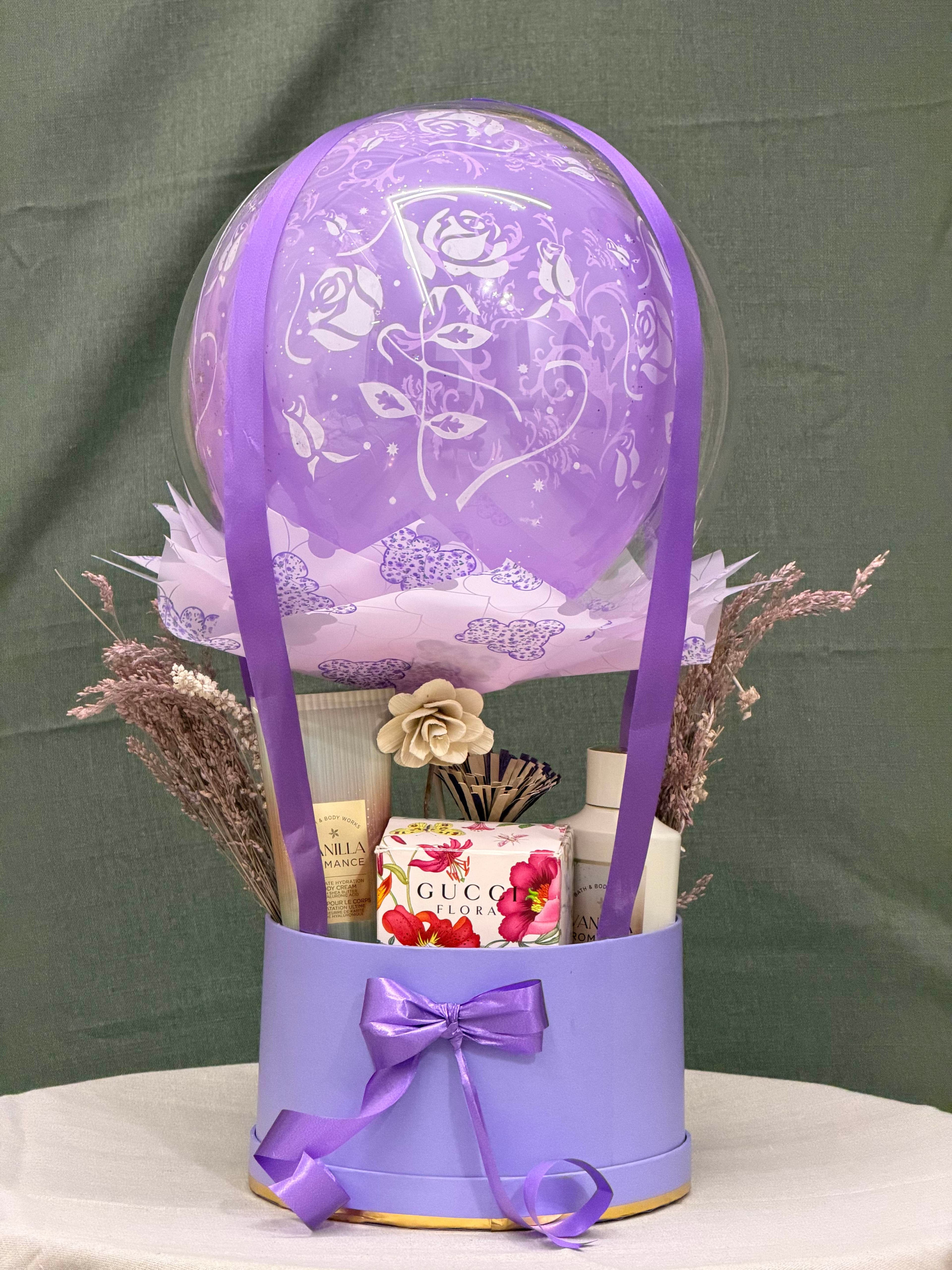 A purple hot-air balloon themed gift set containing various body care and fragrance products, including a Bath & Body Works Vanilla Romance cream and a Gucci Flora perfume/lotion. The set is presented in a round purple box adorned with a matching ribbon bow, and features dried decorative flowers.