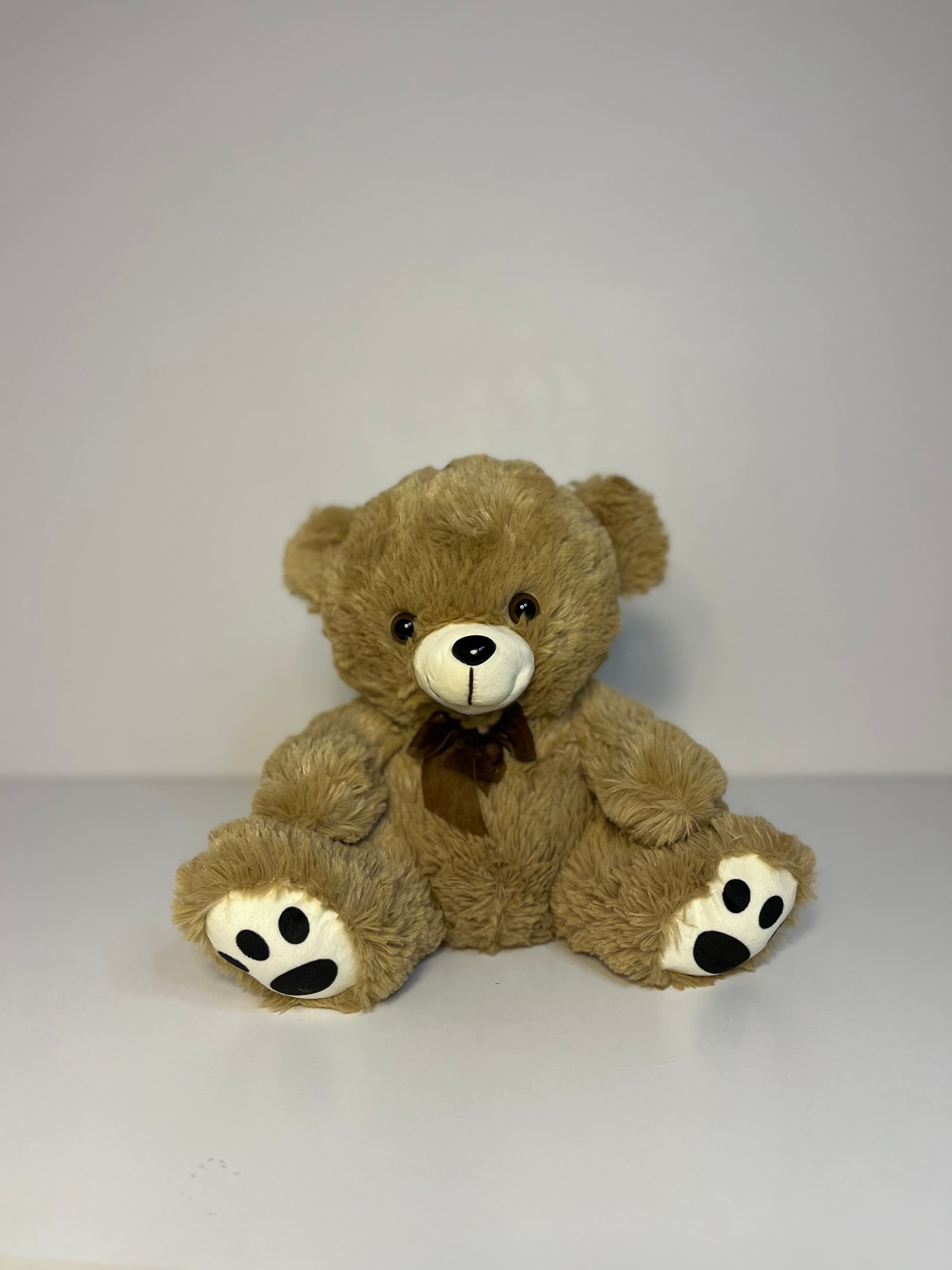 A cuddly brown teddy bear made of soft, fluffy plush material, featuring black button eyes, a stitched black nose and mouth, and white paw pads with black embroidered paw print details, adorned with a small brown ribbon bow around its neck.