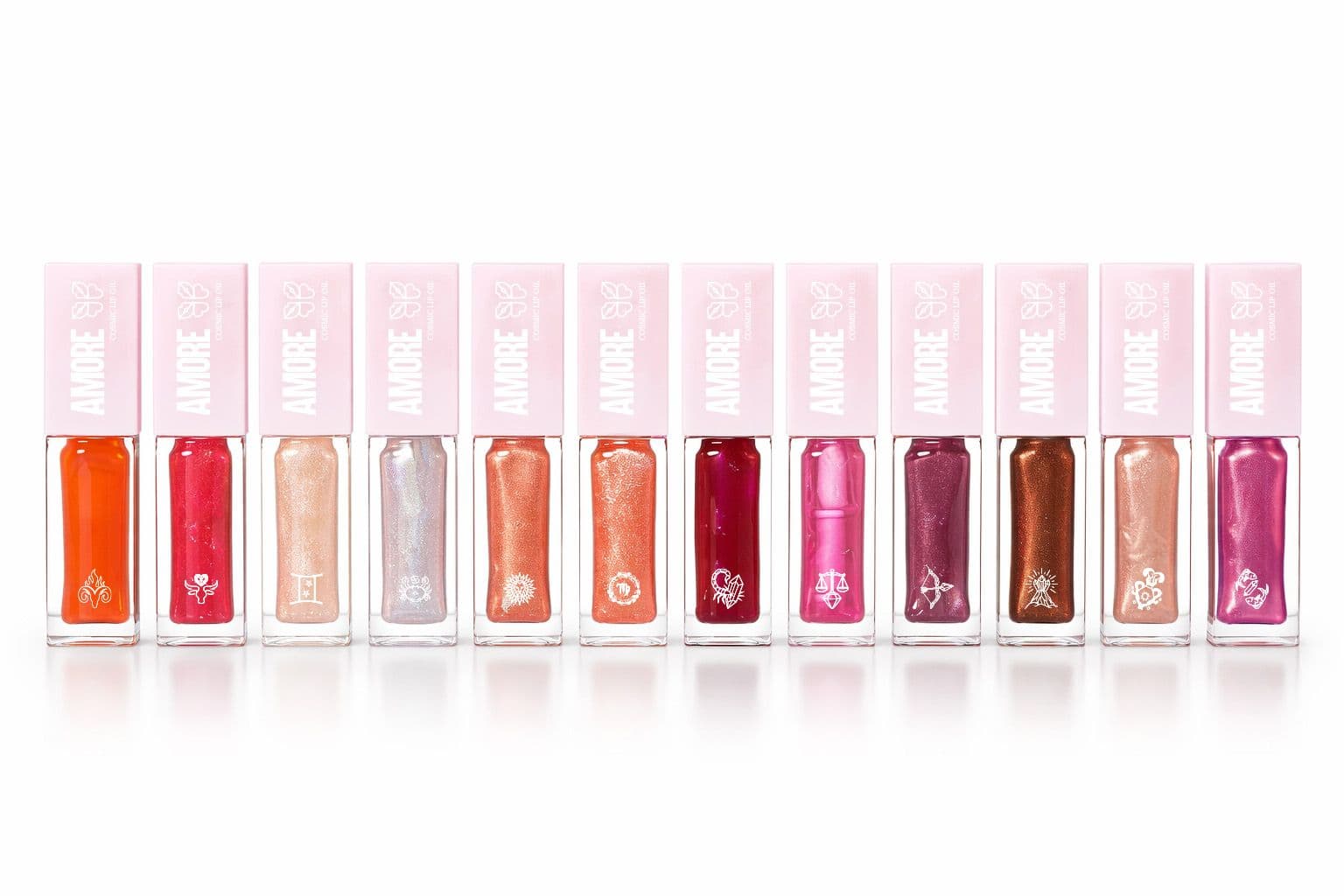 A collection of twelve Amore Cosmic Lip Oils, presented in clear rectangular tubes with light pink caps and featuring various shimmery shades, each with a unique zodiac symbol embossed at its base.