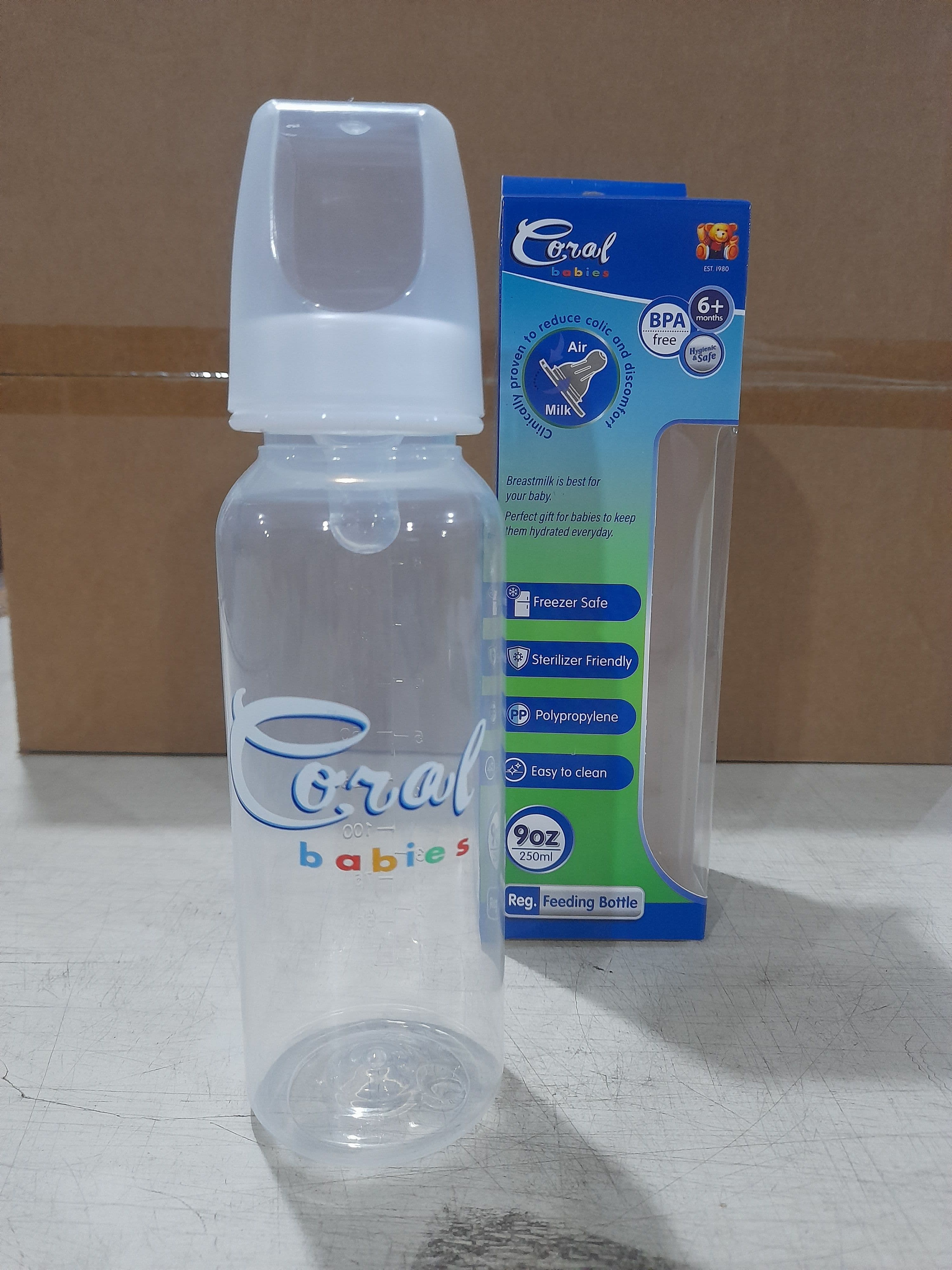 Clear plastic baby feeding bottle with a white nipple and cap, branded 'Coral babies', shown next to its retail packaging highlighting features like anti-colic, freezer safe, and sterilizer friendly.